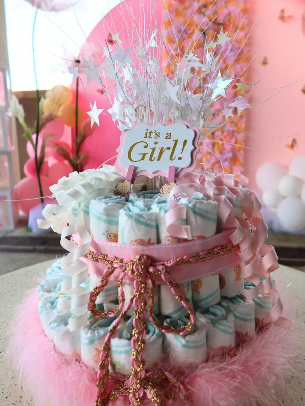 A baby shower diaper cake with a "it's a girl!" topper, adorned with pink ribbons and decorations.