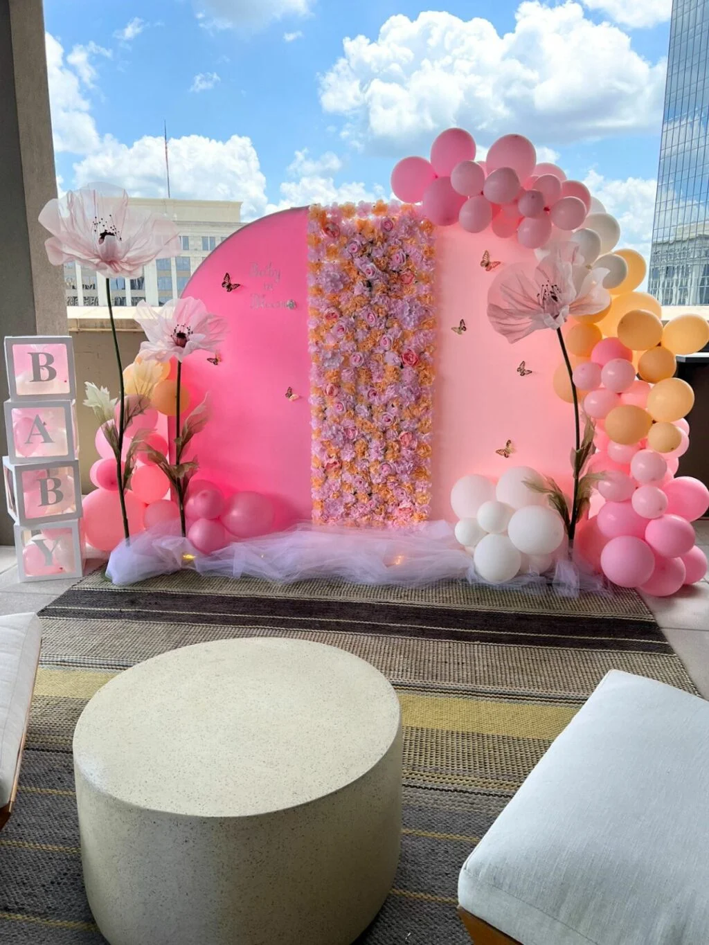 Baby shower balloon decorating services with pink and white balloon arch, floral backdrop, and 'baby' sign set against an urban skyline .