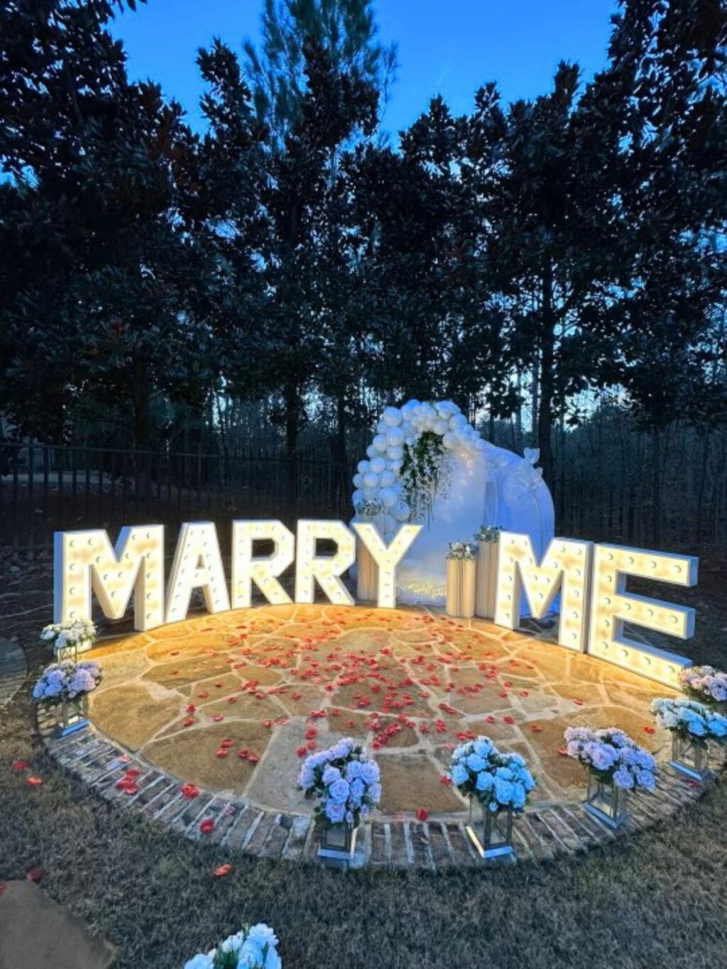 Event decor services for an outdoor marriage proposal setup with illuminated "marry me" sign and floral decorations at dusk.