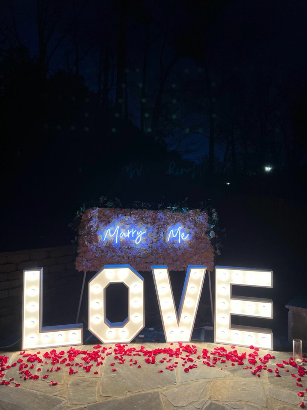 A nighttime marriage proposal setup with illuminated marquee letters spelling 'love' sign and 'marry me' available for event rentals.