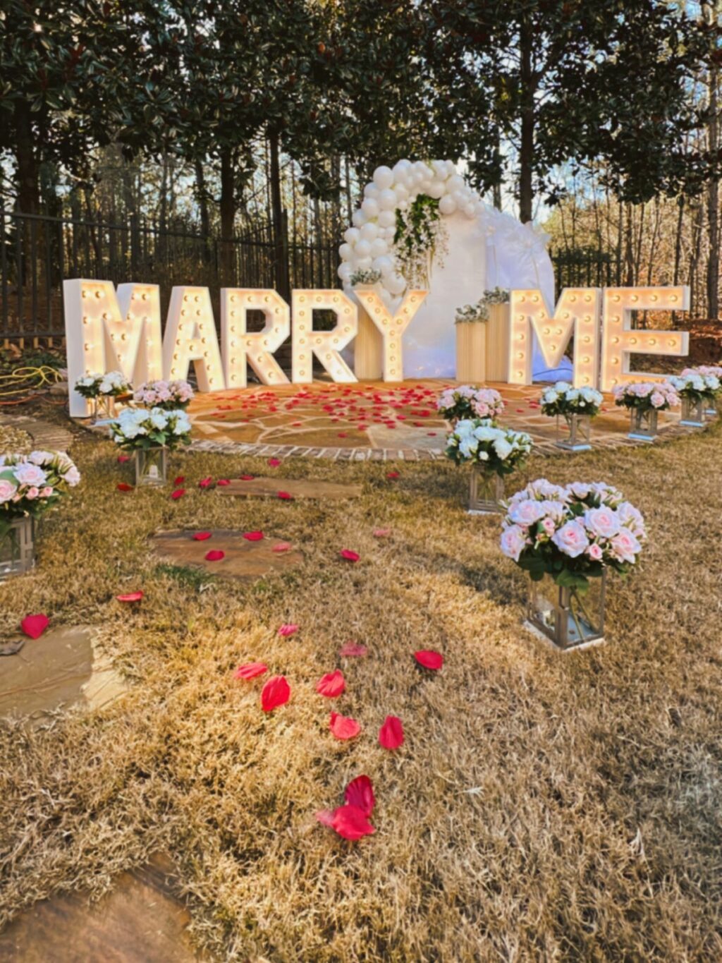 Outdoor marriage proposal setup with large "marry me" letters, flowers, and rose petals.