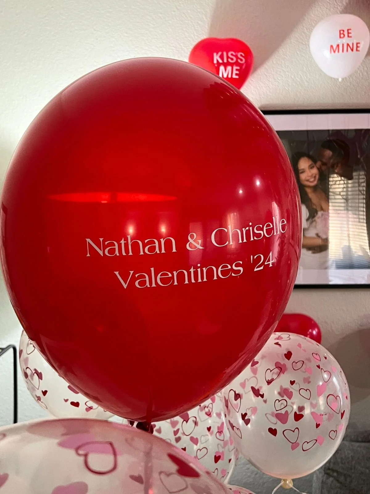 A personalized red balloon with "nathan & chriselle valentines '24" written on it, surrounded by white balloons with heart patterns, with a framed photo and valentine-themed decorations in the background by White Cloud Event Decorators.