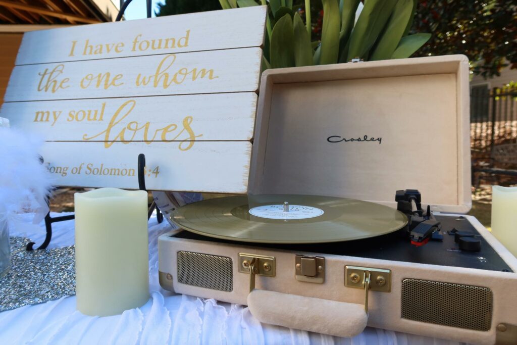 A portable turntable with a vinyl record playing next to a decorative sign featuring a quote from song of solomon 3:4 and a lit candle.