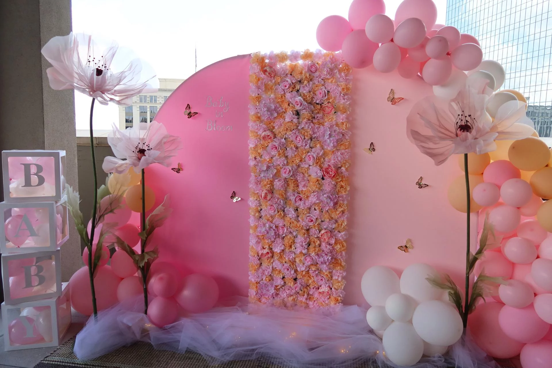 A pink-themed baby shower backdrop with balloon decorations and a floral arrangement.