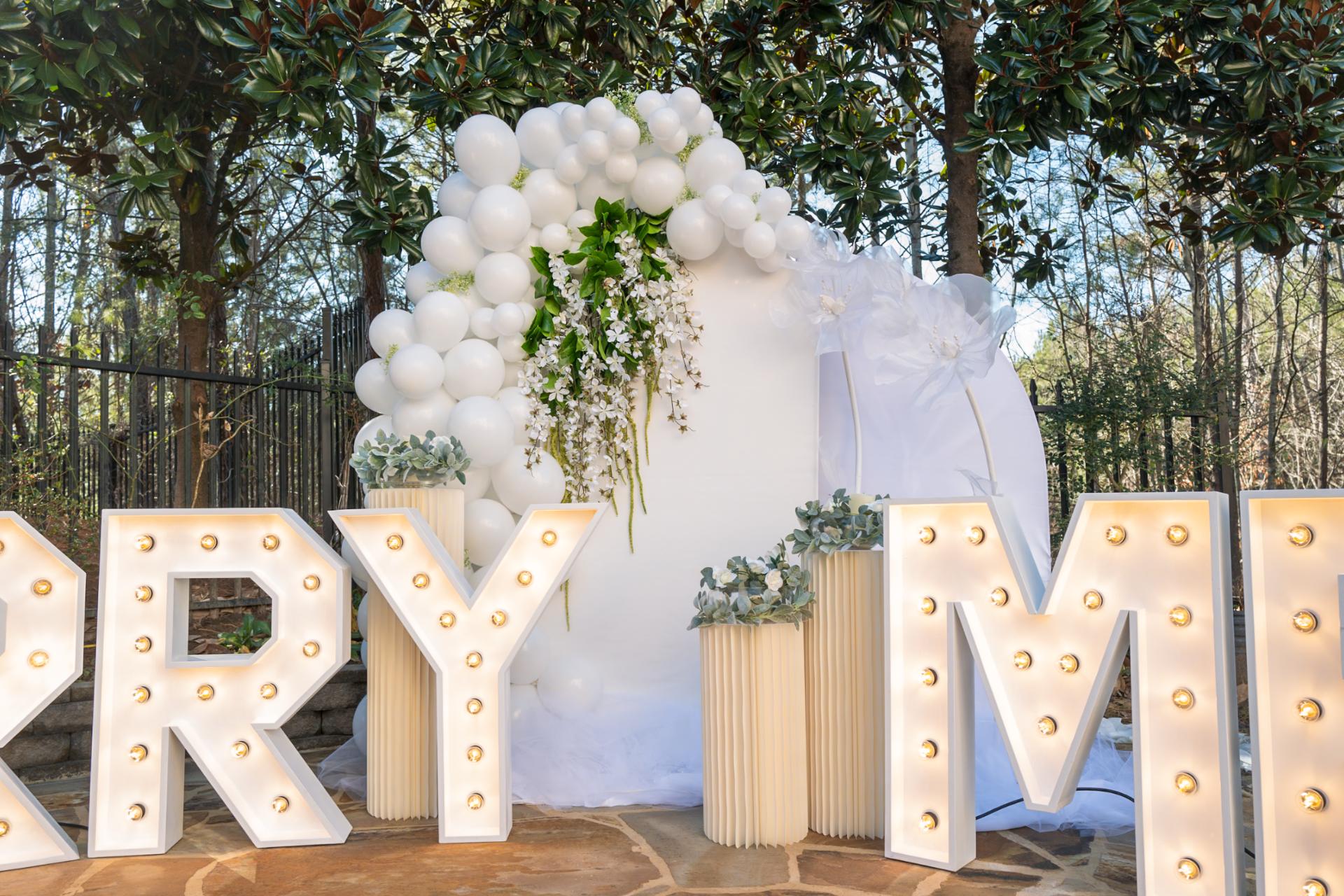 A wedding decoration setup featuring large marquee letters spelling "marry me" in front of a balloon and floral archway.