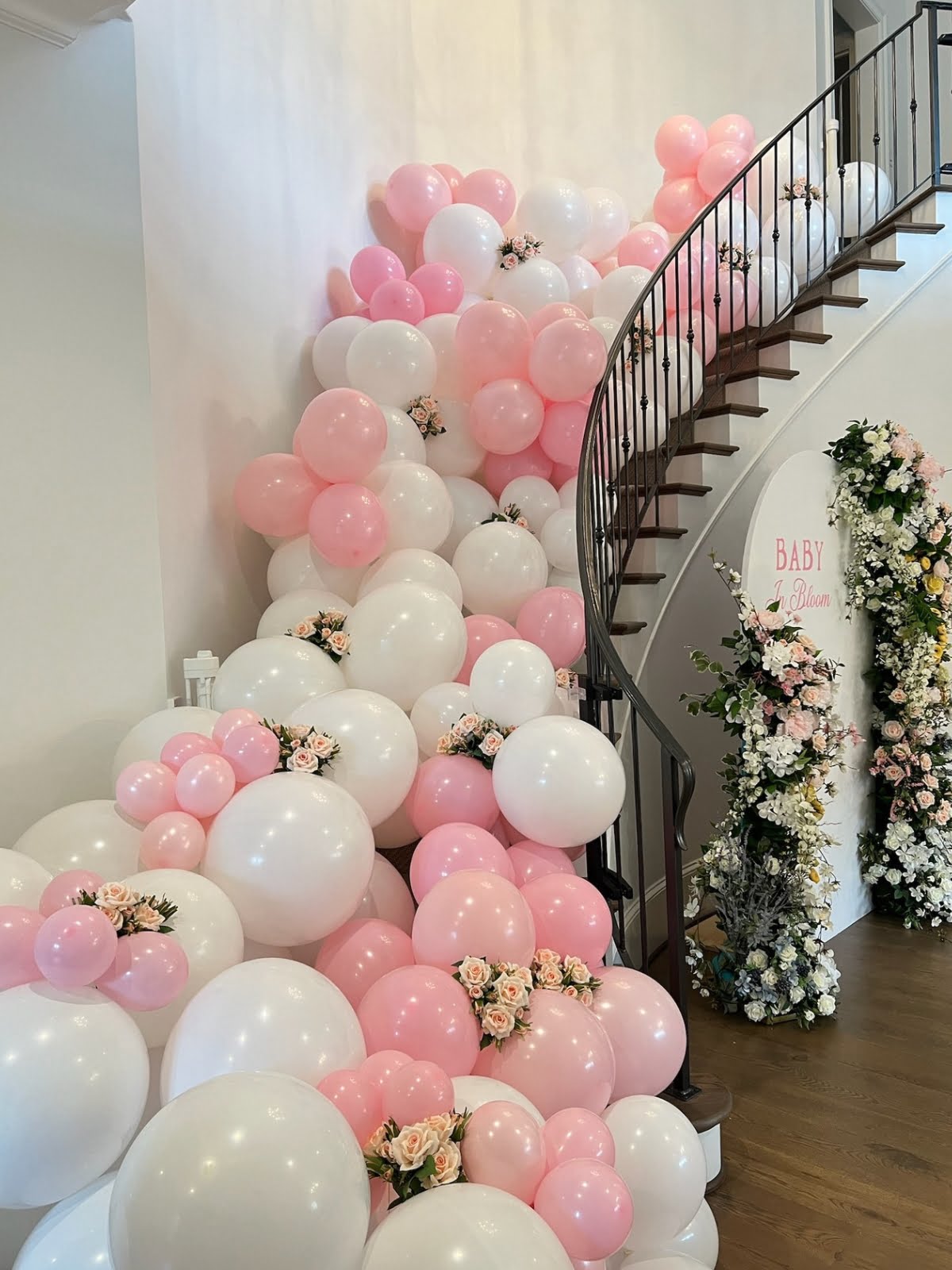 A staircase adorned by event decorators with white and pink balloons cascading down, complemented by flower arrangements interspersed among them. A charming flower arch and a sign reading "BABY" near the staircase add a finishing touch crafted by expert event planners.