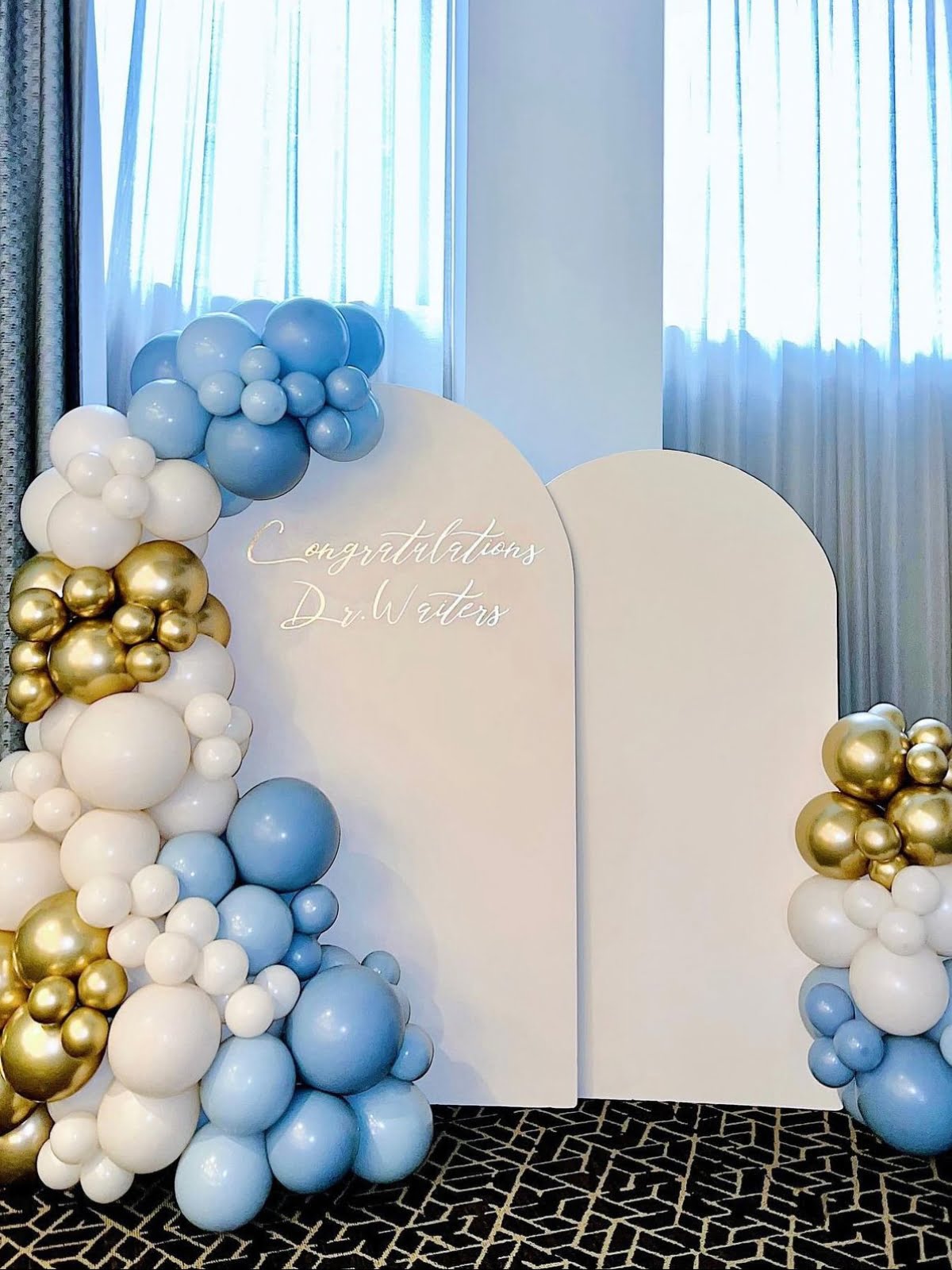 Decorative white arch with blue, white, and gold balloons arranged on either side. The text "Congratulations Dr. Waters" is displayed on the arch, expertly crafted by top event planners to celebrate this special occasion.