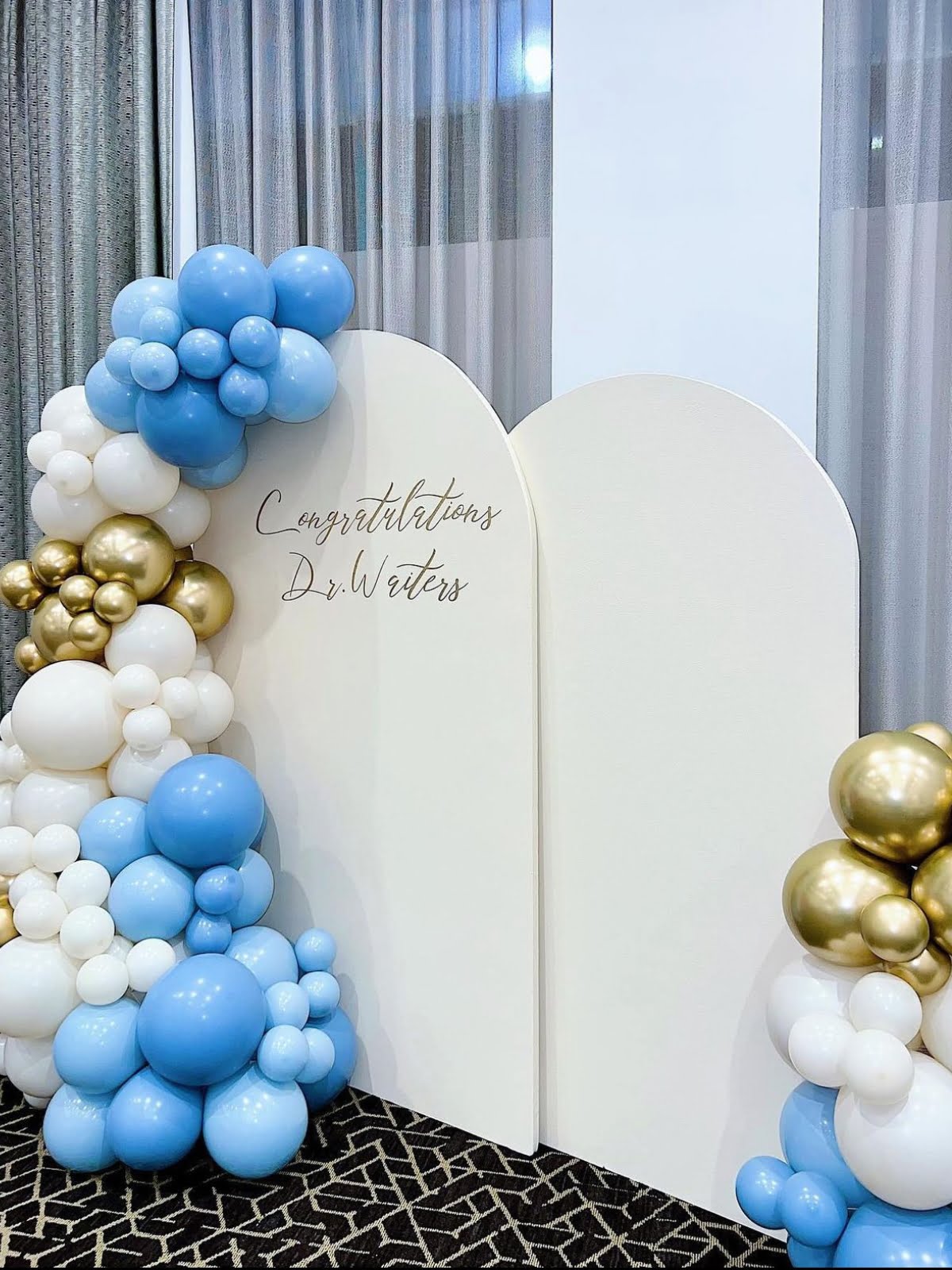 A decorative backdrop with blue, white, and gold balloons, arranged by expert event planners, features a message reading "Congratulations Dr. Waters" on a carpeted floor.