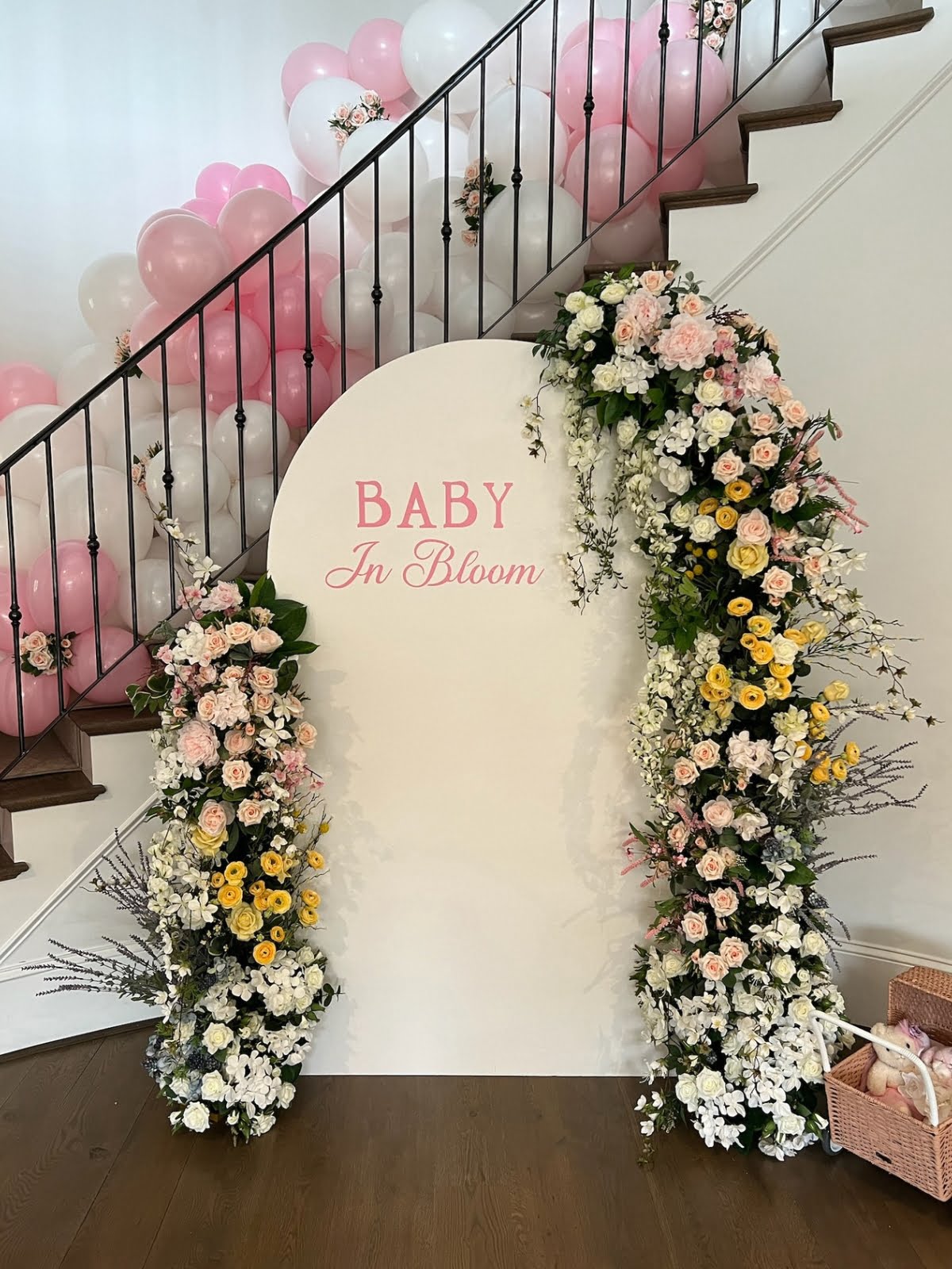 A white sign reading "Baby In Bloom," adorned with floral arrangements, is placed near a staircase with pink and white balloons tied along the railing, showcasing the expertise of talented event planners.