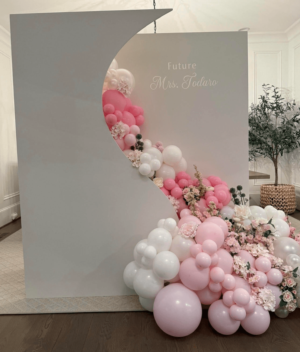 A white decorative wall with “Future Mrs. Todaro” text, featuring a curved opening filled with pink and white balloons and flowers, showcases our balloon decorating services in a modern indoor space.